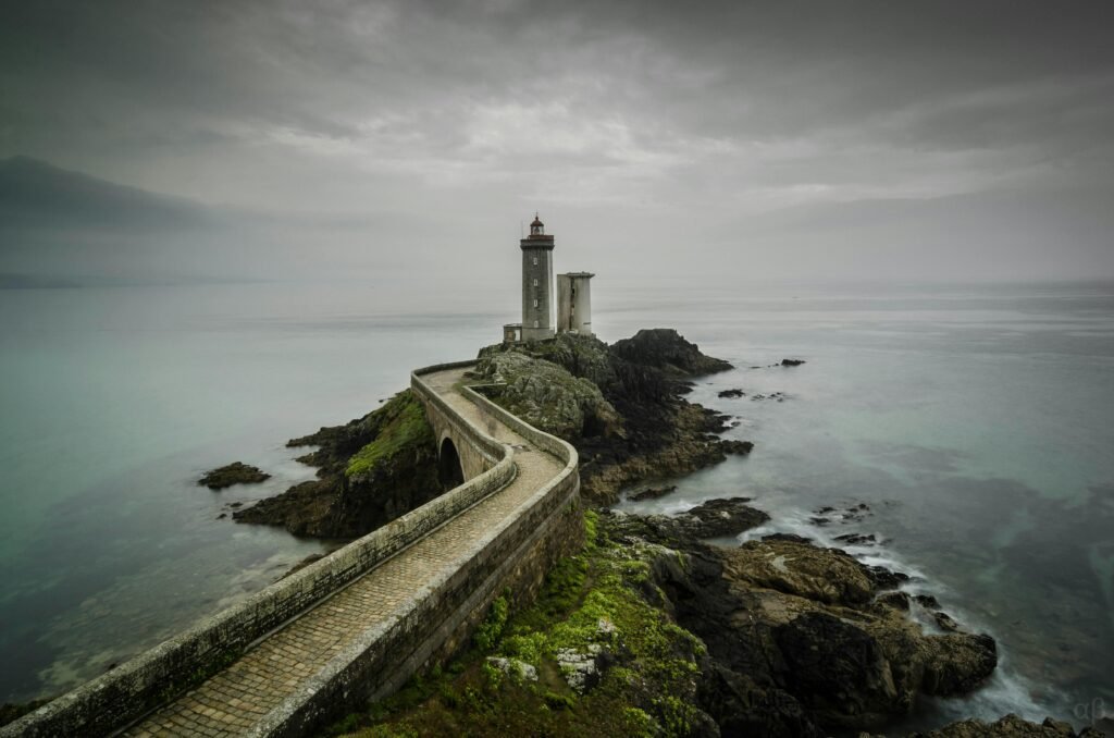 Accueil A scenic view of Le Phare du Petit Minou lighthouse on a cloudy day, situated on a rocky coast in Bretagne, France.